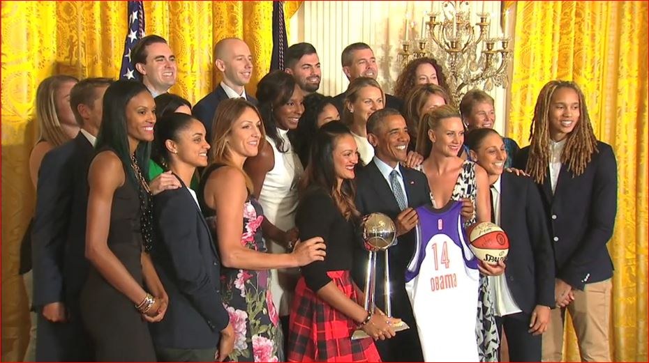 Präsident Obama und First Lady Michelle Obama posieren mit dem Frauen-Basketball-Team im Oval Office des Weißen Hauses, halten einen Basketball, Pokal und lächeln in der Nähe einer Flagge, Vorhänge und einer Kerzenleuchter.
