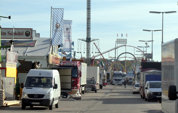 Vollgestellte Straße mit Lastwagen, Autos, Fußgängern, Laternen, Plakaten, einem Turm, einem Vergnügungsgerät, Bäumen und einem bewölkten Himmel.