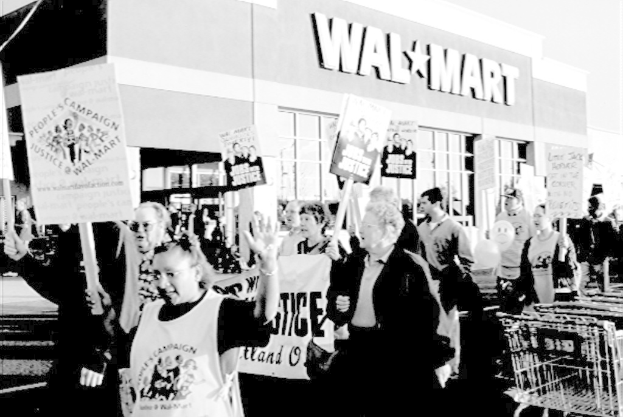 Ein belebter Stadt-Einkaufszentrum mit einem "City Mall Now Open"-Schild, umgeben von Gebäuden, Namensschildern, Geländern, Pflanzen, Bäumen, Strommasten mit Drähten und einem bewölkten Himmel.