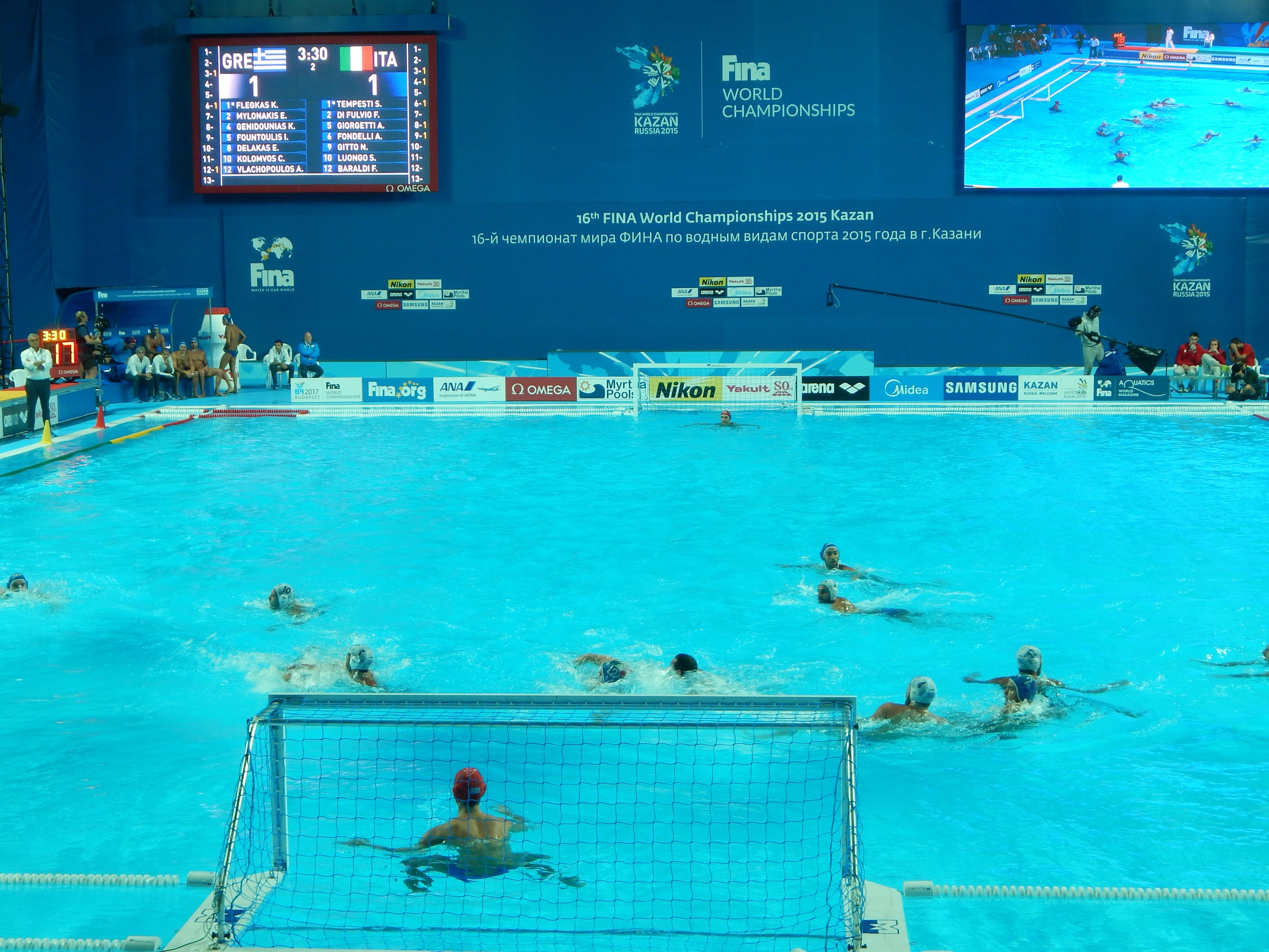 Wasserballspieler bei den 2016 FINA-Weltmeisterschaften in Kazan im Pool gegeneinander antretend, mit Zuschauern auf der linken Seite und einem Werbeschild im Hintergrund.