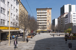 Ein belebter Stadtplatz mit Menschen, die spazieren gehen, auf Bänken sitzen oder stehen, umgeben von Bäumen, Gebäuden und einem Karussell im Hintergrund unter einem sichtbaren Himmel.