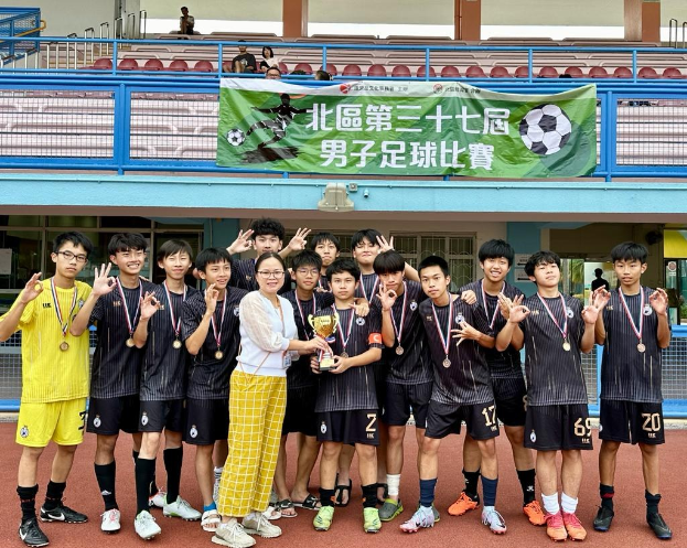 Gruppe junger Männer in Fußballtrikots auf einem Feld mit einem Pokal und Medaillen, im Hintergrund ein 'Yokohama U-16 Boys Soccer Team'-Plakat.