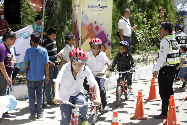 Kinder fahren mit Fahrrädern eine Straße entlang, die mit Verkehrskegeln markiert ist, einige tragen Helme, andere stehen daneben, mit einem Banner, Bäumen und Gebäuden im Hintergrund.