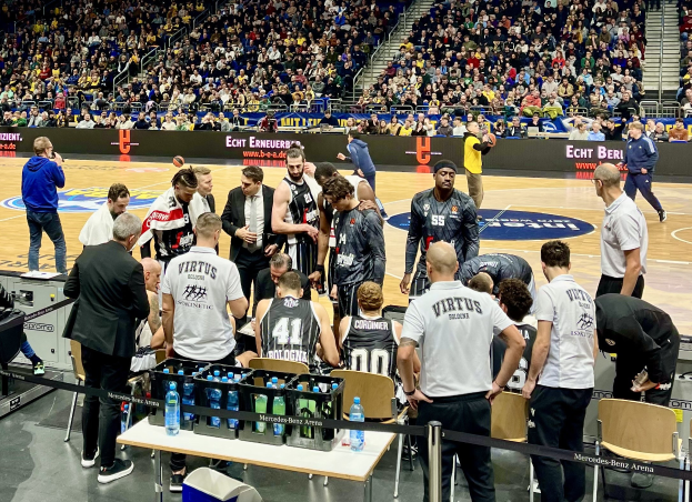 Gruppe von Männern auf einem Basketballfeld während eines Spiels zwischen Virtus Berlin und Echt Berlin, mit einer Zuschauermenge in der Nähe, einem Fotografen mit Equipment links und Informationsschildern im Hintergrund.