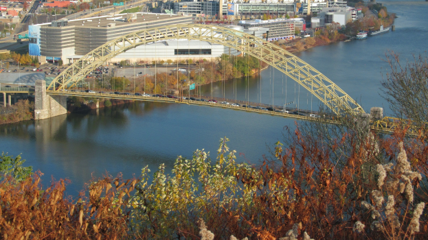 Eine Brücke spannt einen Fluss mit fahrenden Autos darauf, vor einer Kulisse aus Bäumen und Gebäuden.
