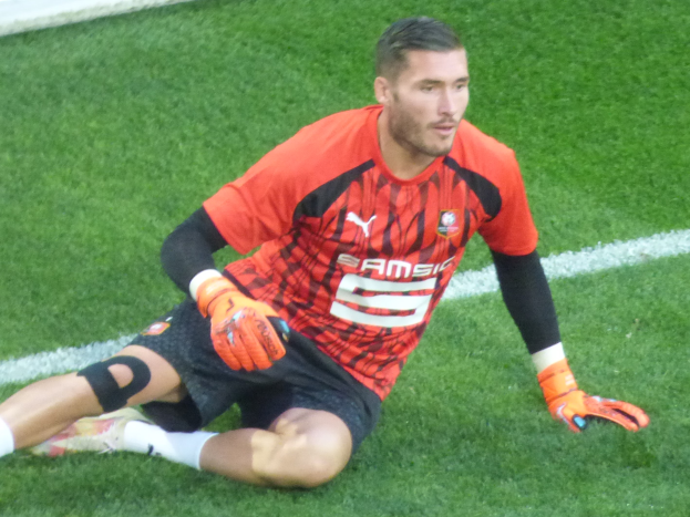 Ein Fußballspieler in rotem T-Shirt, schwarzen Shorts, weißen Socken und orangefarbenen Baseballhandschuhen sitzt auf dem Boden mit Händen auf den Knien.