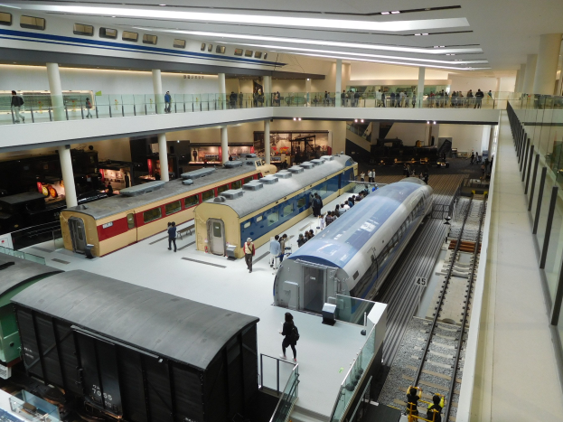 Ein großer Museumsraum mit verschiedenen Zügen in unterschiedlichen Größen und Farben, bei dem Menschen die Ausstellungsstücke vom Boden, Geländern und Säulen unter Deckenlampen betrachten.