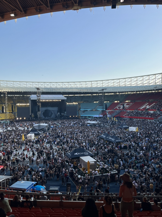 Große Menschenmenge sitzt und steht in einem Stadion mit Zelten, Geländern und Beleuchtung von oben, was auf ein Konzert in der O2 Arena in London hindeutet.