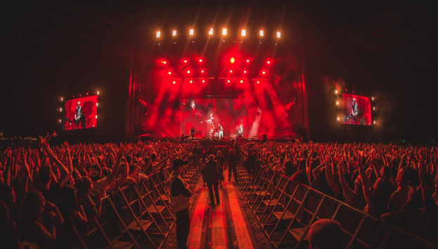 Eine große Menschenmenge steht vor einer hell erleuchteten Bühne mit Geländern, auf der Musiker spielen, während andere das Konzert gegen einen dunklen Hintergrund beobachten.