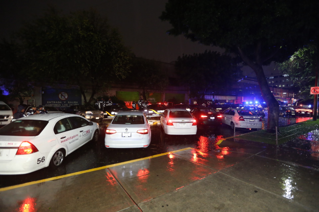 Eine Gruppe von Polizeiwagen auf einem Parkplatz bei Nacht, mit einem Schild, Gras, Bäumen, Gebäuden, einer Fahne, Lichtern und einem bewölkten Himmel.