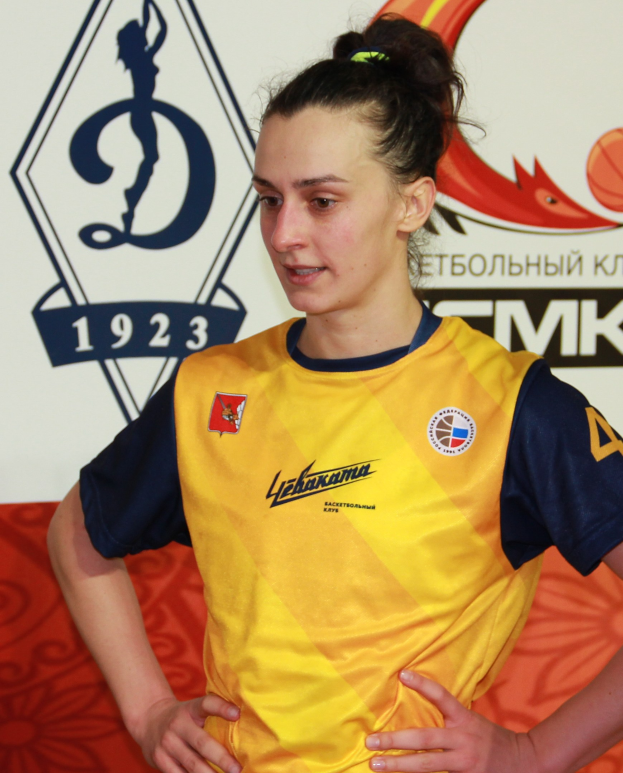 Eine Handballerin in gelb-blauem Trikot steht mit Händen in den Hüften, entschlossen schauend, vor einem Banner mit Text und Logo.