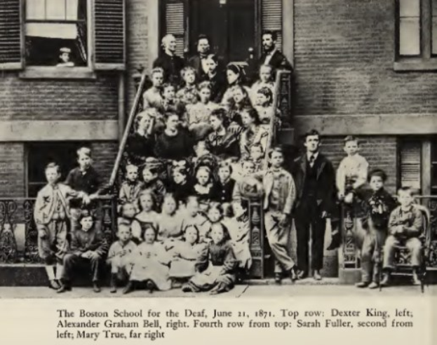 Gruppe von Menschen, die vor einem Gebäude mit der Aufschrift "The Boston School for the Deaf, June 21, 1871" stehen und sitzen