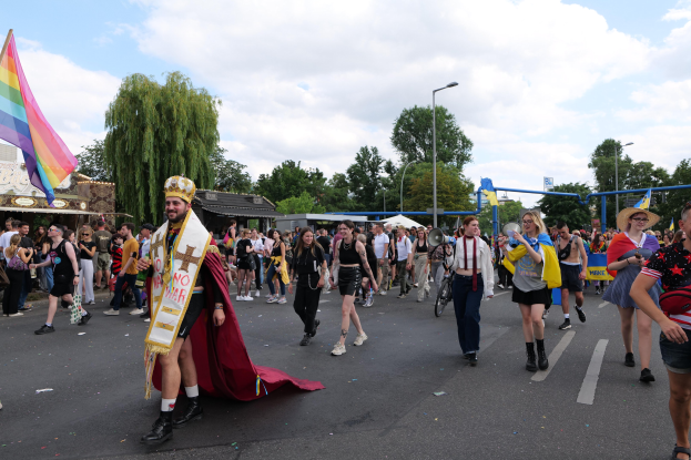 Eine Gruppe von Menschen marschiert bei der Gay Pride Parade 2018 mit Musikinstrumenten und einer Regenbogenflagge, im Hintergrund sind Laternenpfähle, Bäume, Hütten und ein bewölkter Himmel zu sehen.