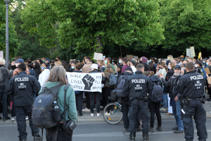 Eine große Gruppe von Menschen steht an der Straße, einige halten Schilder, mit einem Fahrrad im Vordergrund und Bäumen und einem Pfahl im Hintergrund, bei einer Black Lives Matter Demonstration in Berlin.