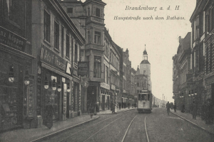 Ein Schwarz-Weiß-Foto einer Stadtstraße in Brandenburg, Deutschland, mit einer Straßenbahn auf Schienen, Menschen auf dem Bürgersteig, Gebäuden mit Fenstern, Schildern und Straßenlaternen unter einem bewölkten Himmel.