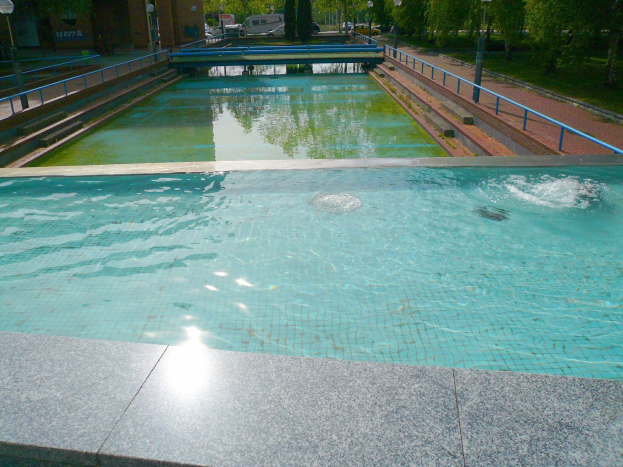 Großes Schwimmbad in einem Park, umgeben von Geländern, Laternen, Bäumen und Fahrzeugen, mit Gebäuden und einem klaren blauen Himmel im Hintergrund.