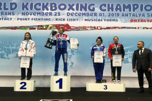 Eine Gruppe von fünf Personen auf einem Podium stehend, jede mit einem Pokal und Urkunden in der Hand, mit einem Banner im Hintergrund mit der Aufschrift "Welt-Kickbox-Meisterschaft Antalya 2019".