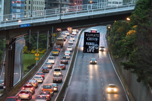 Eine belebte Stadtstraße mit starkem Verkehr, eine Brücke im Hintergrund und ein Banner mit Text in der Mitte der Straße, flankiert von Bäumen, Polen, Lampen, Schildern und Gebäuden auf der rechten Seite.