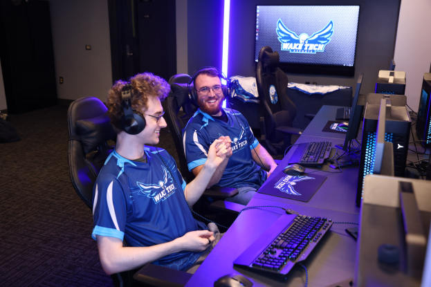 Zwei Männer in blauen T-Shirts und Kopfhörern sitzen an einem Schreibtisch mit Computer-Monitoren, Tastaturen und anderem Equipment und spielen ein Spiel, mit Taschen auf dem Boden links und einem Bildschirm und Lichtern im Hintergrund.