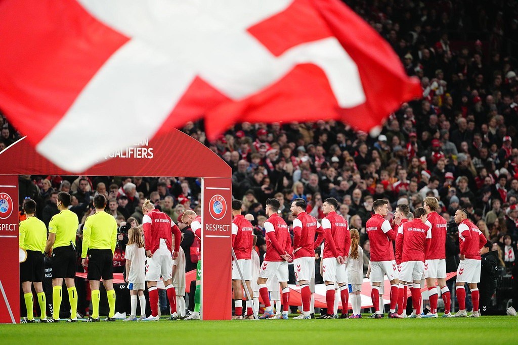 Gruppe von Menschen auf einem Fußballfeld mit einer roten und weißen Flagge im Vordergrund, einem Bogen mit der Aufschrift "Bayern München vs. Bayern München Wett-Tipps & Vorschau" im Hintergrund und einer großen Menge im Stadion.