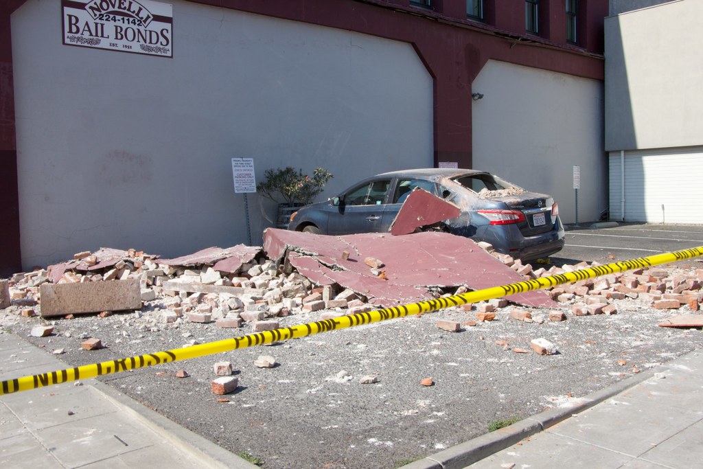 Eine zerbrochene Ziegelwand liegt auf dem Boden mit verstreuten roten Ziegeln, einem geparkten Auto, einer Topfpflanze und einem Gebäude mit einem "Kautionsgeschäft"-Schild im Hintergrund.