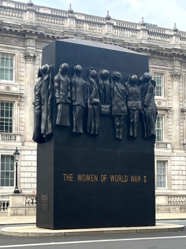 Das Frauen-Denkmal des Zweiten Weltkriegs in London, eine große schwarze Struktur mit eingraviertem Text, steht umgeben von einer Straße, Laternenmasten, Geländern und einem Gebäude mit Fenstern im Hintergrund unter einem bewölkten Himmel.