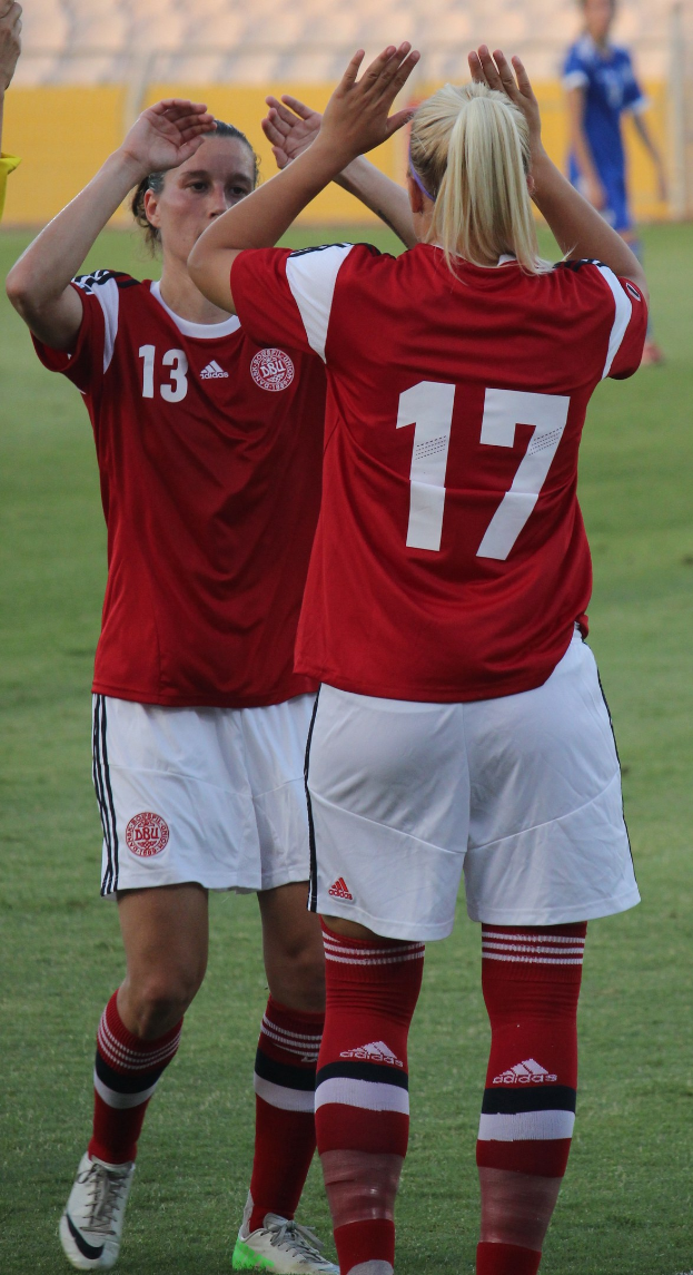 Zwei Frauen in roten T-Shirts und weißen Shorts klatschen auf einem Fußballfeld, mit einer Person in einem blauen Kleid und einem gelben Brett im Hintergrund sowie Stühlen.