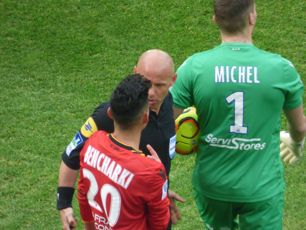 Zwei Fußballspieler und ein Schiedsrichter auf einem grasbedeckten Feld, wobei einer der Spieler einen Ball hält.