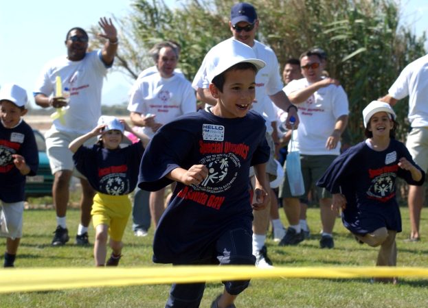 Kinder bei einem Lauf auf einem grünen Feld mit einem gelben Band im Vordergrund, einige tragen Kappen und Schutzbrillen, andere halten Gegenstände, vor einem klaren blauen Himmel mit Bäumen im Hintergrund.