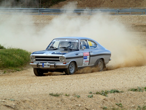 Ein Auto fährt auf einer Schotterstraße mit Rauch aus dem Auspuff, umgeben von Gras auf der linken Seite und einem Geländer im Hintergrund, mit Passagieren im Auto und Text und Zahlen auf dem Auto.