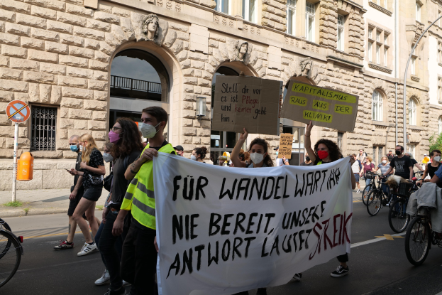 Eine Gruppe von Menschen, die bei einer Demonstration durch die Straße marschieren, einige halten Schilder und andere fahren Fahrräder, im Hintergrund ein Gebäude mit Rundbogenfenstern, Säulen und Skulpturen.