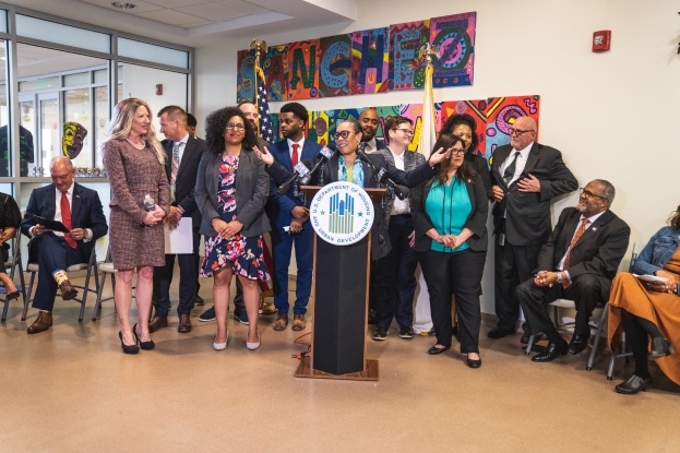 Gruppe von Menschen vor einem Podium mit einem Mikrofon und einer Tafel mit dem Text "Sancho City Councilman Joins City Council", mit sitzenden und stehenden Teilnehmern, Flaggen, einem Feuerlöscher und Deckenlampen im Hintergrund.