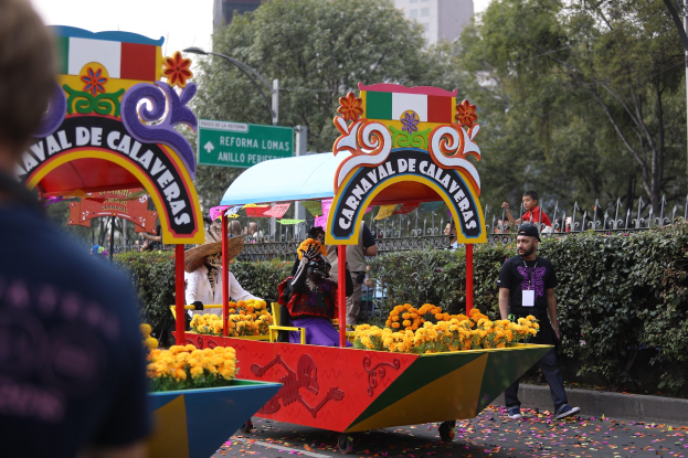 Eine Gruppe von Menschen auf einem blumengeschmückten Festwagen bei einem Umzug, umgeben von einem Metallzaun, Schildern, einer Straßenlaterne, einer Metallabsperrung, Pflanzen, Bäumen und einem Gebäude mit Fenstern unter einem bewölkten Himmel.