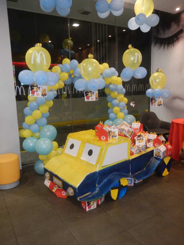 Eine McDonald's-Geburtstagsfeier mit einem gelben und blauen Lastwagen, der mit Ballons geschmückt ist, einem roten Tisch mit Gegenständen, Hockern, einer Glaswand und Deckenlampen in einer festlichen Umgebung.