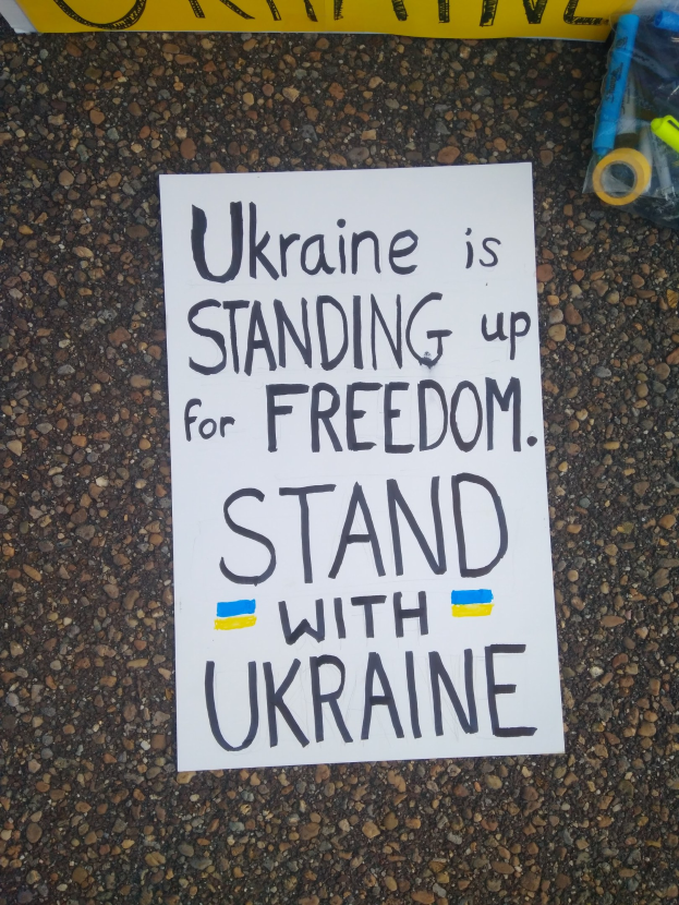 Ein weißer Papierbogen mit den Worten "Ukraine steht für Freiheit auf, stellt Euch mit Ukraine" liegt vor einer gelben Fahne auf dem Boden, rechts daneben eine Plastikabdeckung mit verschiedenen Gegenständen.