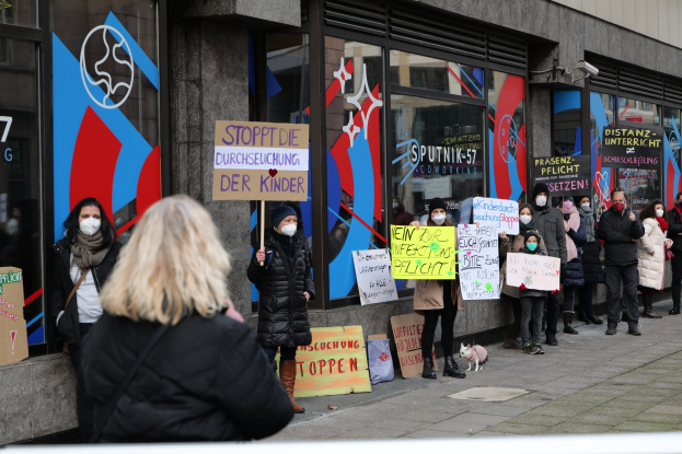 Gruppe von maskierten Menschen mit Schildern und Plakaten vor einem Glasfrontgebäude, bei der Kameras und ein Hund anwesend sind, protestiert gegen ein Schulmaskenverbot.