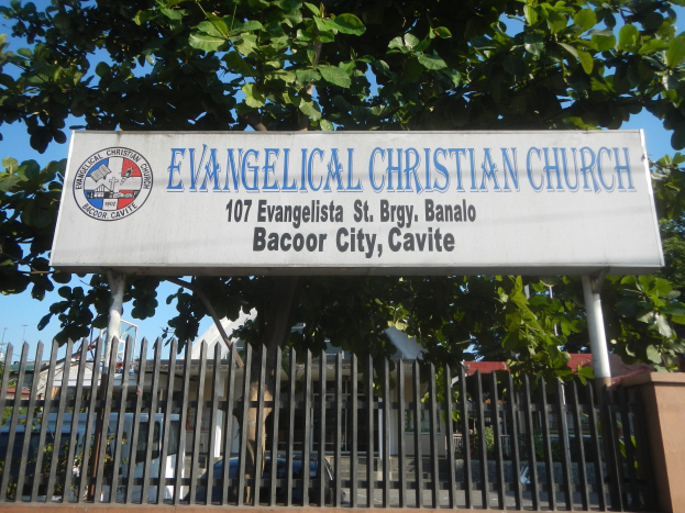 Evangelische Kirche in Bacoor City, Cavite, mit einem Schild, einem Metallzaun, einer Wand, Bäumen, Fahrzeugen, benachbarten Gebäuden, Strommasten und einem bewölkten Himmel.