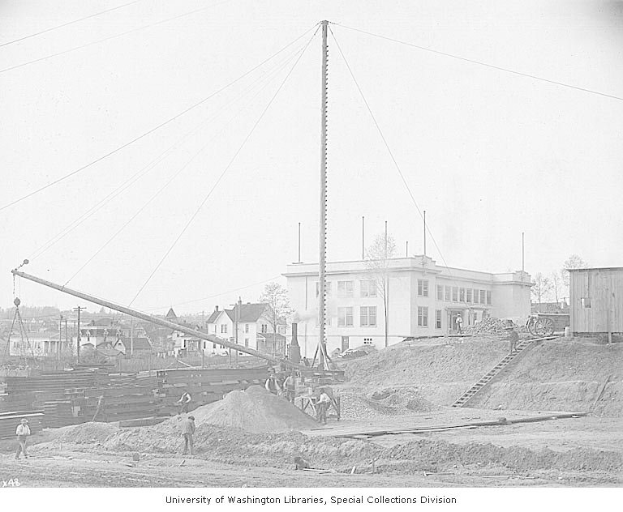 Schwarzes und weißes Foto einer Baustelle mit einem großen Gebäude im Hintergrund, Arbeitern, Baustoffen, Bäumen und einem bewölkten Himmel, beschriftet mit "Bau des neuen Bibliotheksgebäudes an der University of Washington Libraries, Special Collections Division."