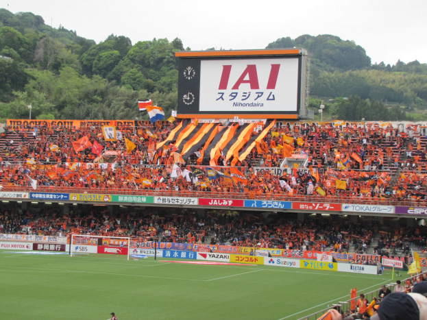 Ein Fussballspiel auf einem grünen Feld mit einem Tor, umgeben von einer grossen Zuschauermenge in den Stadionrängen, Schildern, Fahnen, einem grossen Bildschirm, Bäumen und einem klaren blauen Himmel.