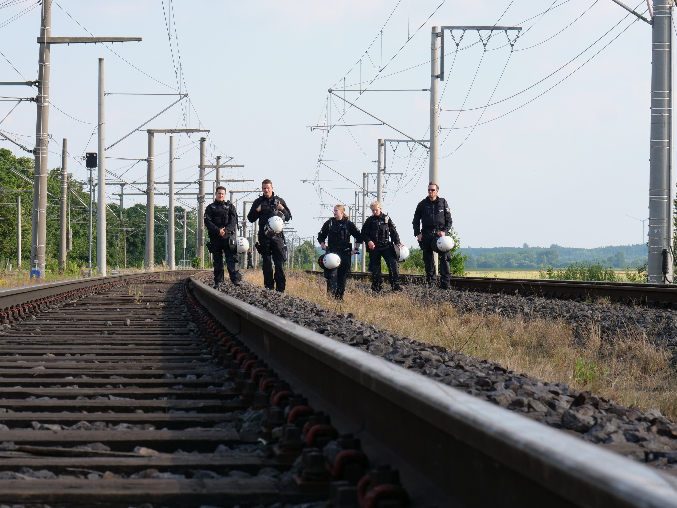 Eine Gruppe von Polizisten geht auf den Schienen, jeder hält einen Helm in der Hand, mit Strommasten, Bäumen und einem klaren blauen Himmel im Hintergrund.