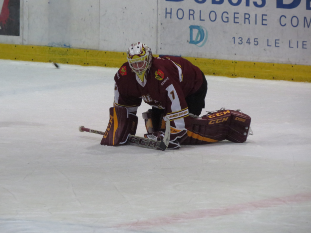 Eishockeyspieler in rot-gelber Uniform, der einen Schuss auf dem Eis abwehrt, trägt Helm, Handschuhe und Knieprotektoren und hält einen Eishockeyschläger, mit einer Wand und Text im Hintergrund.