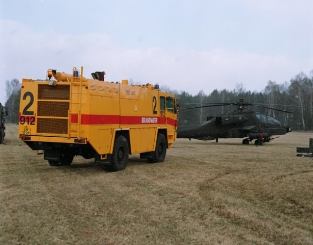 Ein Hubschrauber mit einem Seil fliegt über eine grüne Landschaft und löscht ein entferntes Waldfeuer, das von Rauch und Bäumen umgeben ist.