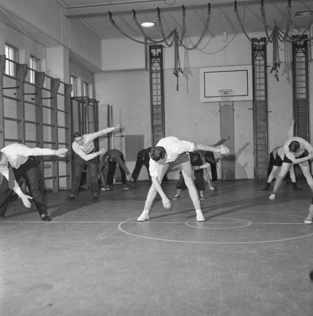 Black and white image of a group performing gymnastics in a gymnasium with a wall, windows on the left, and ceiling lights in the background.