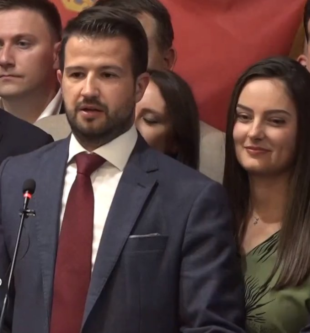 Ein Mann im Anzug spricht an einem Rednerpult mit Mikrofon zu einer Menge hinter einem Banner, wahrscheinlich über ein Regierungsverbot von Social Media.