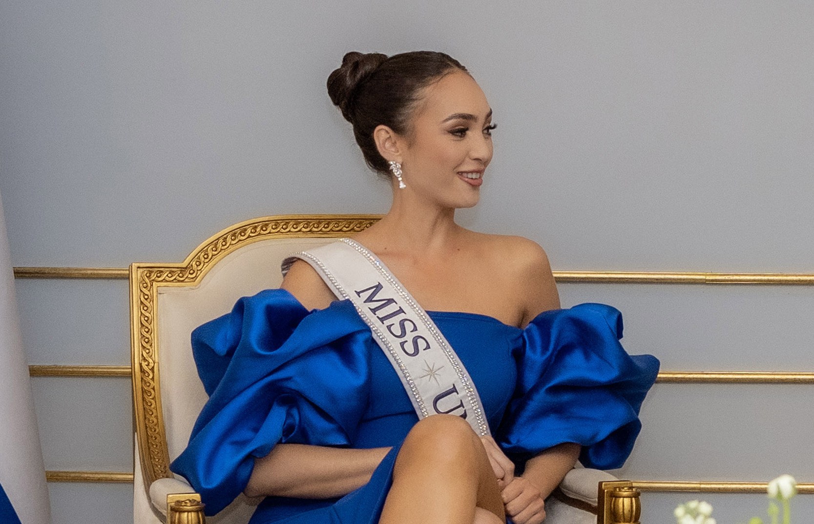 Eine Frau in einem blauen Kleid trägt eine "Miss Universe 2019" Schärpe und sitzt lächelnd auf einem Stuhl umgeben von Blumen mit einer Wand im Hintergrund.