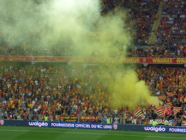 Große Menschenmenge in einem Fußballstadion mit Fahnen, Bannern und Rauch in der Luft.