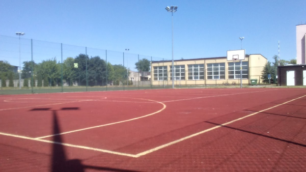 Korb eines Basketballplatzes umgeben von Gebäuden, Straßenlaternen, einem Mülleimer, einem Zaun, Bäumen und einem klaren blauen Himmel.