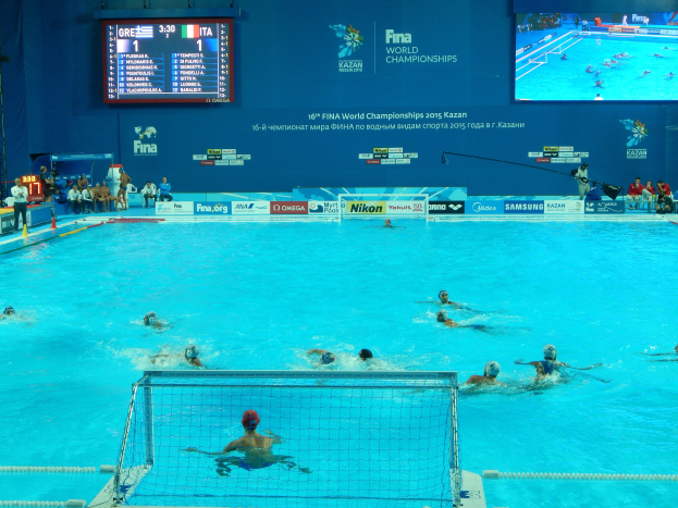 Wasserballspieler bei den 2016 FINA World Championships in Kazan im Pool gegeneinander antretend, mit Zuschauern auf der linken Seite und einem Anzeigebrett mit Event-Branding im Hintergrund.