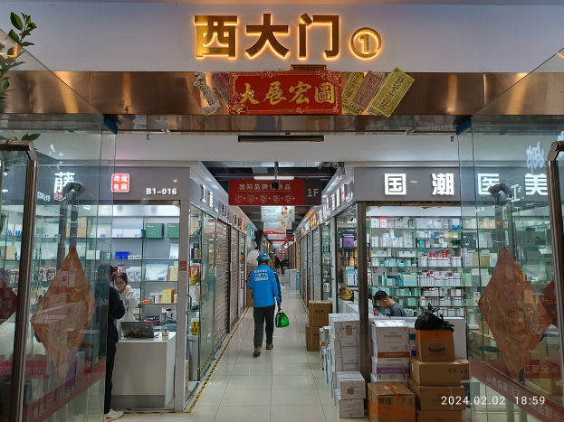 Innenraum einer chinesischen Apotheke mit Glaswänden, Regalen voller Artikel, verstreuten Kartons, einer Pflanze links und einem Schild oben.