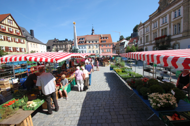 Ein belebter Markt im alten Stadtkern von Heidelberg mit Menschen, die spazieren gehen, auf Bänken sitzen und in der Nähe von Zelten stehen, mit Gemüsekörben auf Tischen, Gebäuden mit Fenstern, Bäumen und einem klaren blauen Himmel.